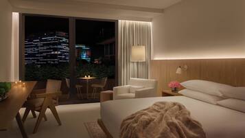 A modern hotel room with a large bed, a dining area with a table and chairs, a nightstand with a lamp, and a view of the cityscape at night.