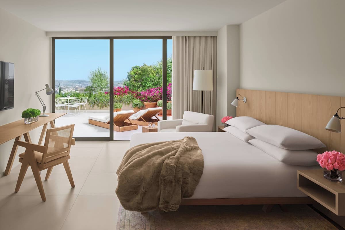 Een moderne hotelkamer met een groot bed, een bureau, een stoel, en een balkon met zitplaatsen buiten en uitzicht op groen.