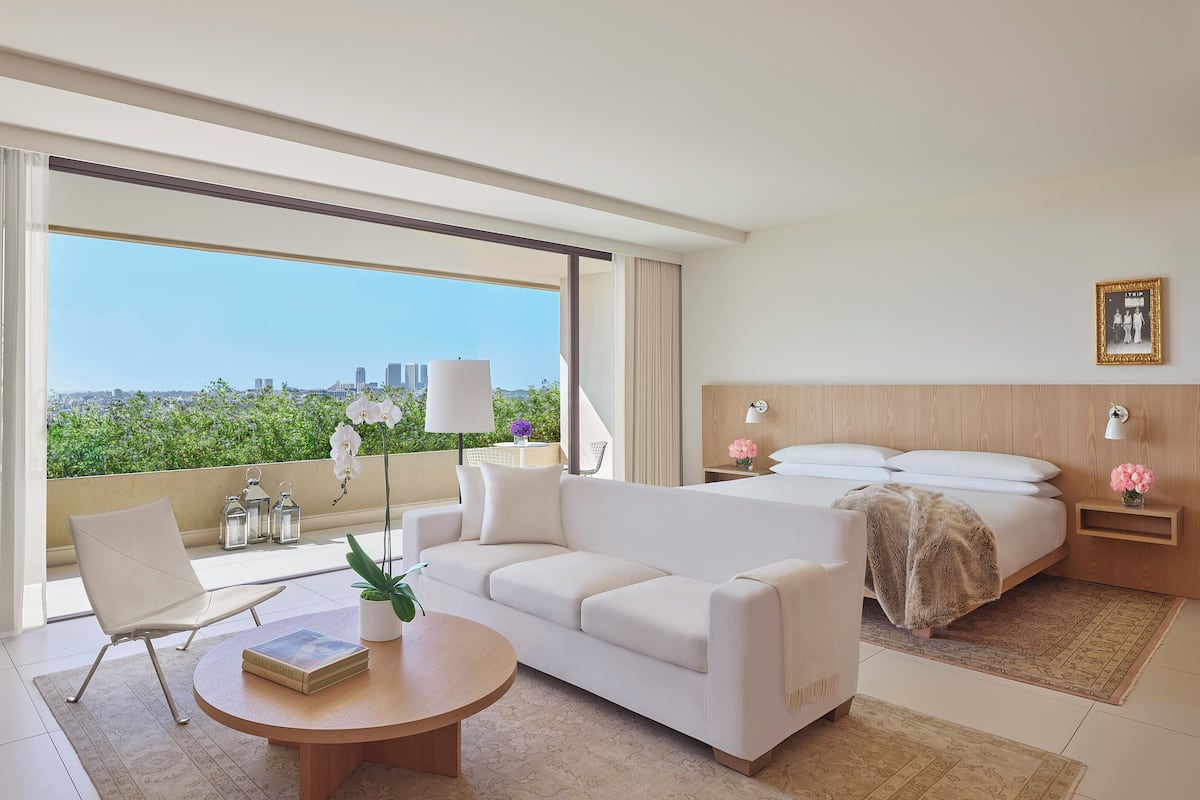 Een moderne hotelkamer met een groot balkon, een bank, een ronde salontafel, een bed en een nachtkastje met lampen.