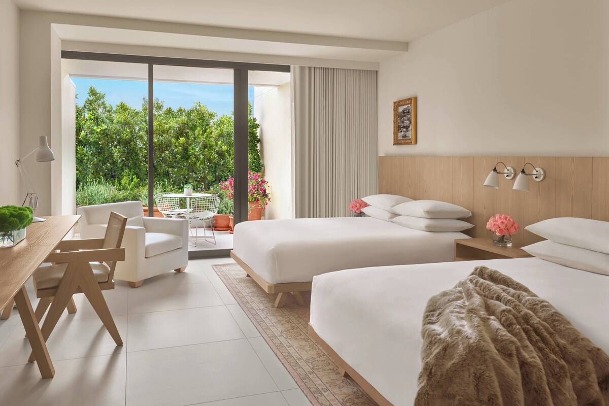 Hotelkamer met twee bedden, een bureau, stoelen, een balkon met uitzicht op groen en bloemen, en een houten hoofdbord.