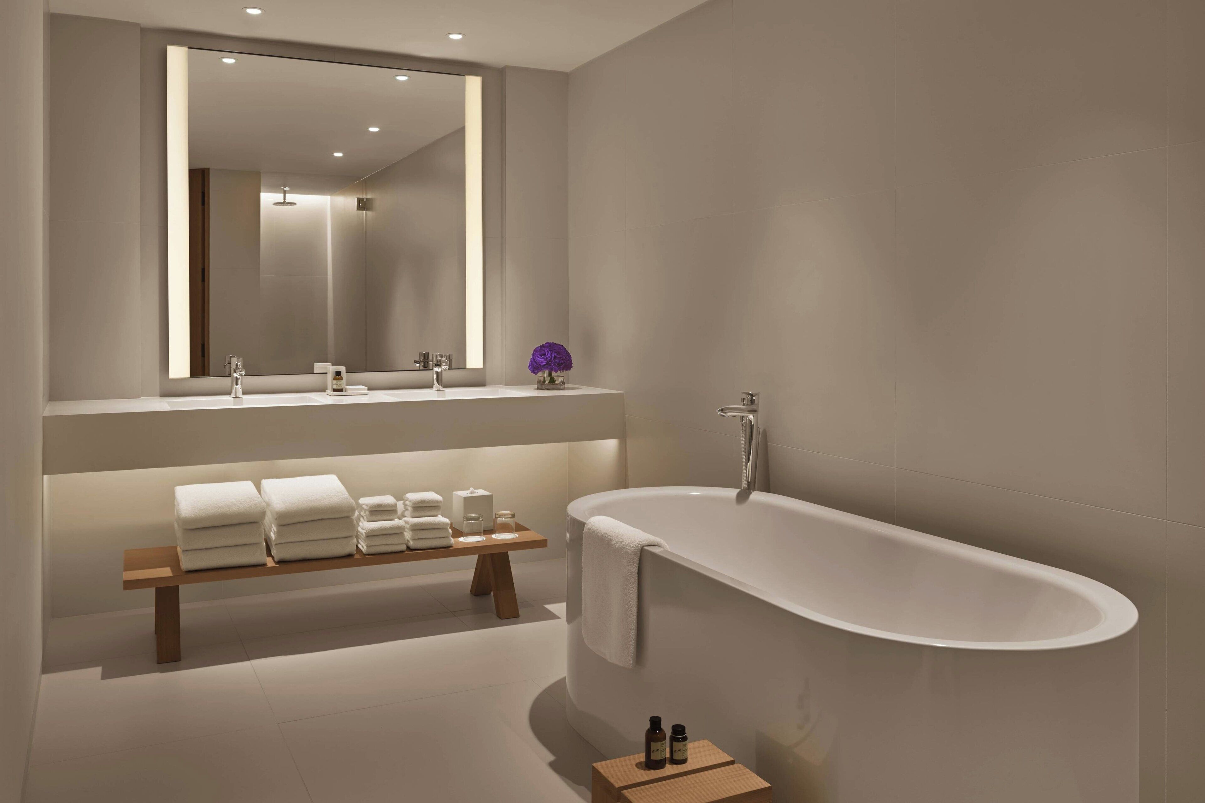 A modern bathroom with a freestanding bathtub, a wooden bench with towels, and a large mirror above the sink.