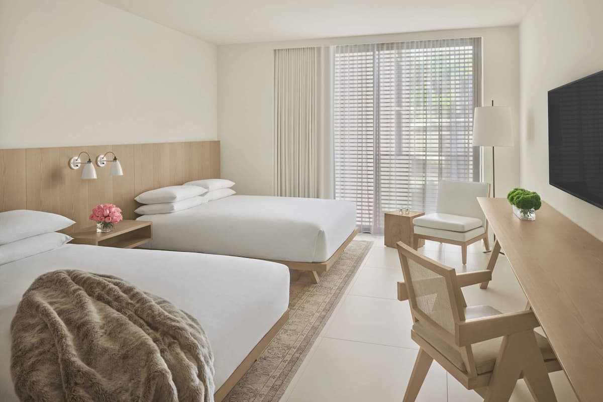 Een hotelkamer met twee bedden, een bureau, een stoel, een televisie en een raam met jaloezieën.
