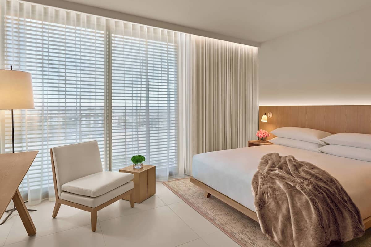 Een moderne hotelkamer met een groot bed, een stoel, een klein tafeltje met een lamp en dunne gordijnen.