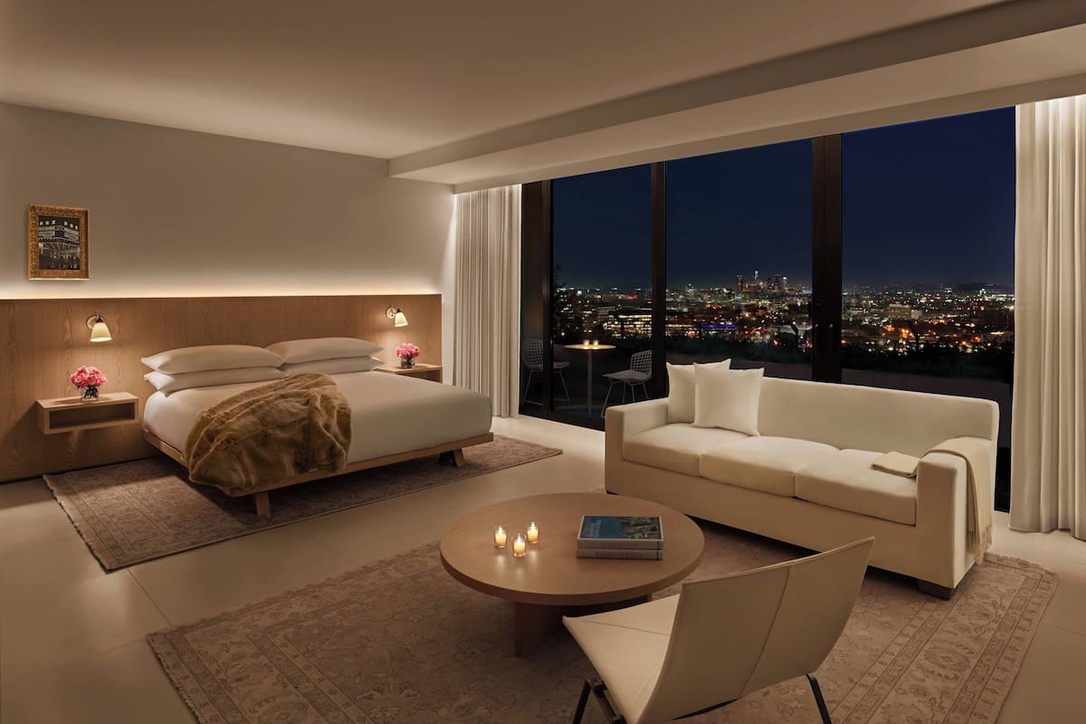 Een moderne hotelkamer met een groot bed, een bank, een bijzettafeltje en uitzicht op een stadsbeeld bij nacht.