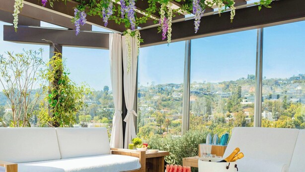 Un espace extérieur moderne, aménagé avec des canapés blancs et une table basse en bois, entouré d’une végétation luxuriante et de fleurs violettes.