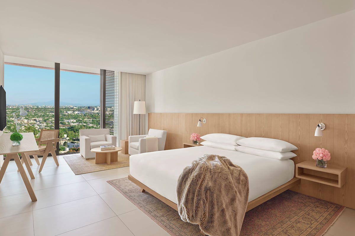 Een moderne hotelkamer met een groot bed, een bureau met stoel, een klein tafeltje en uitzicht op de stad door grote ramen.