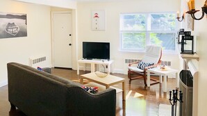 Ein Wohnzimmer mit einer Couch, einem Fernseher, einem Couchtisch und einem Sessel. Es gibt ein Fenster mit Jalousien, eine Tür und eine Wand mit einem Bild.
