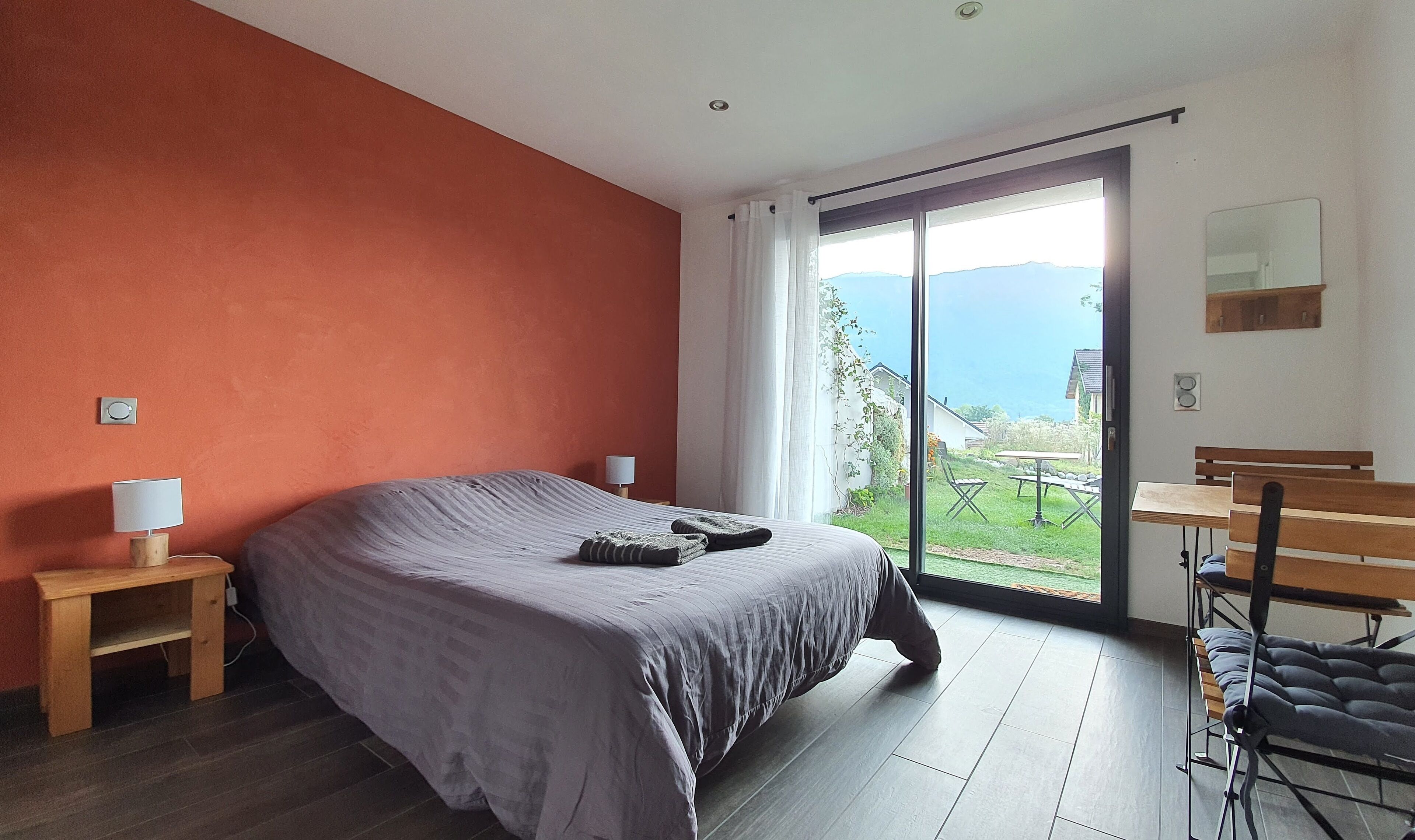 A bedroom with a large bed, a nightstand, a desk with a chair, and a sliding glass door leading to a balcony with a view of the sea.