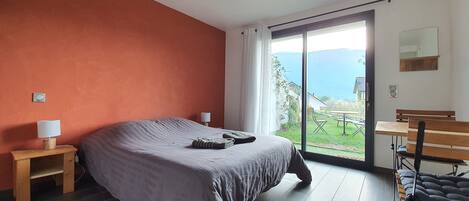 A bedroom with a large bed, a nightstand, a desk with a chair, and a sliding glass door leading to a balcony with a view of the sea.