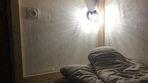 Et lille, svagt oplyst rum med en enkeltseng, en vægmonteret lampe og en synlig stikkontakt.