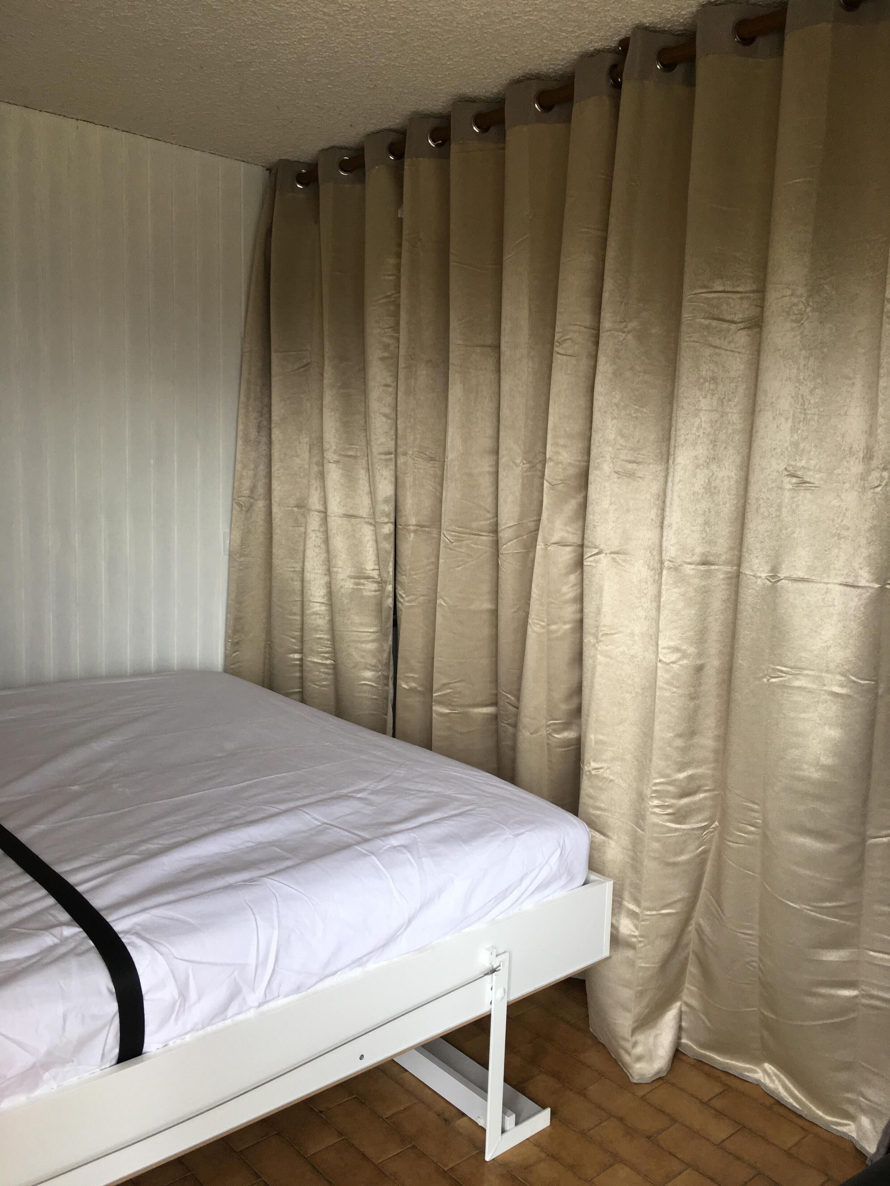 Una cama con colchón blanco y detalles negros, ubicada bajo cortinas color beige.