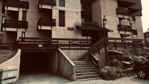 En bygning med flere etager med skilt, der viser "Le Sanctuaire". Der er en trappe op til en indgang, et lille udendørs område med en bænk og en parkeret bil.