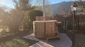 A wooden hot tub with a chimney, surrounded by a garden with a lamp post and a wooden fence.