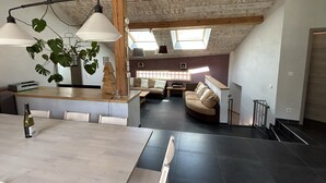 A modern loft with a high ceiling, exposed wooden beams, a dining area with a long table, a kitchen with a bar, and a cozy seating area with a sofa and a coffee table.