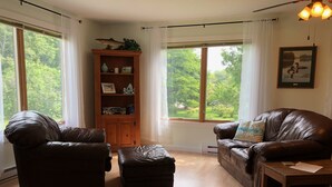 Ein Wohnzimmer mit zwei Ledersofas, einem Holzschrank und einem großen Fenster mit Blick auf Grünflächen.