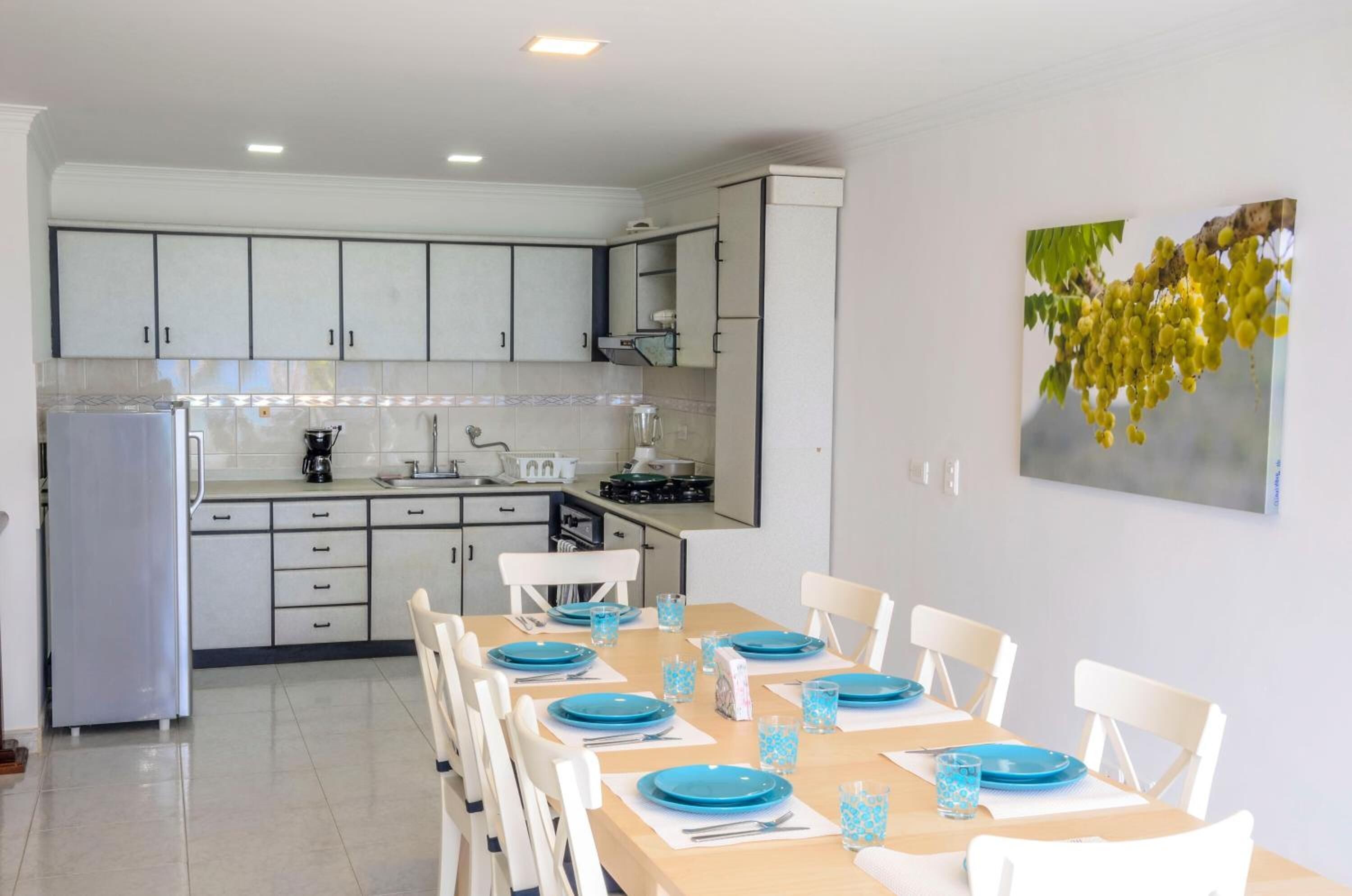 A modern kitchen with white cabinets, a dining table set with blue plates, and a painting of grapes on the wall.