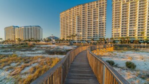 A wooden boardwalk leading to high-rise buildings with balconies, surrounded by sand dunes and sparse vegetation.