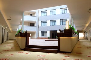 A spacious hotel lobby with a central staircase leading to upper floors, a reception desk, and a view of the building's exterior.