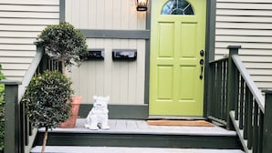 Une porte verte avec une petite fenêtre, une statue de chat blanc et un arbre en pot placé sur les marches.