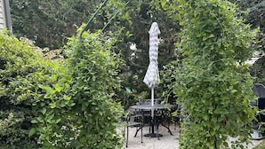 Un chemin pavé de briques mène à un espace patio aménagé avec une table et des chaises, le tout entouré d’une végétation luxuriante.