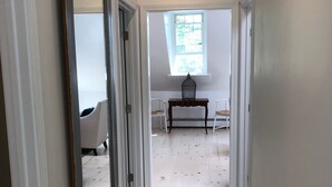 Un couloir avec un plancher en bois, des murs blancs et un grand miroir sur le côté gauche. On aperçoit une fenêtre donnant sur des arbres, ainsi qu’une table et des chaises au fond.