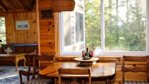 A wooden cabin interior with a dining table, chairs, a sofa, and a large window offering a view of the forest. - Hobby Horse - Rustic Maine Cabin, Ocean Access 22 miles from Acadia Nat'l Park (Surry)