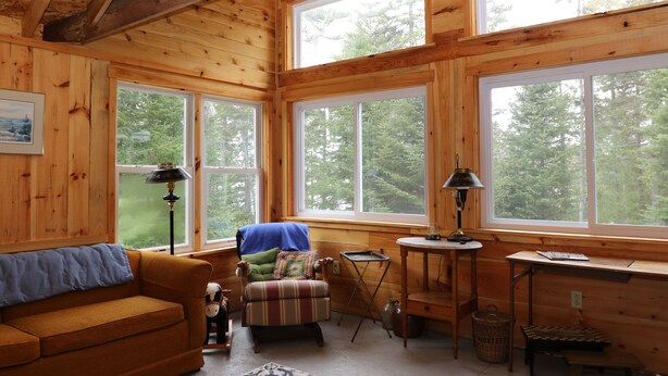 A cozy cabin interior with wooden walls, large windows, a sofa, a chair, a lamp, and a small table. - Hobby Horse - Rustic Maine Cabin, Ocean Access 22 miles from Acadia Nat'l Park (Surry)