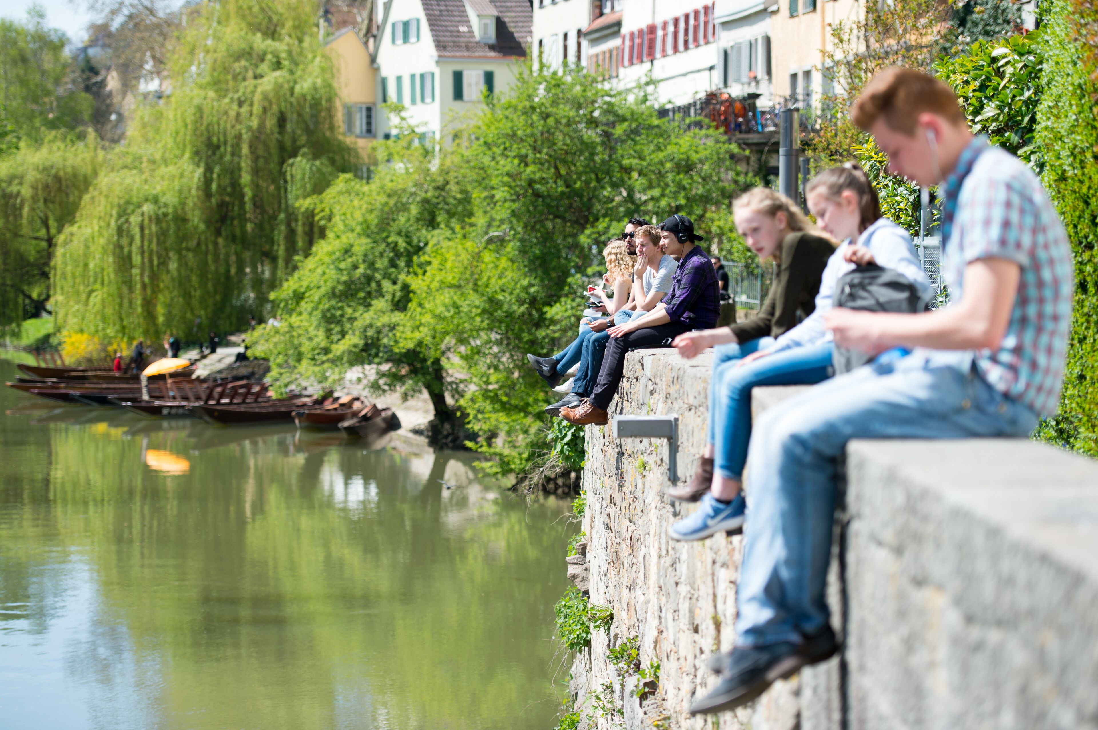 A group of people sitting on a stone wall by a river, with buildings and greenery in the background.