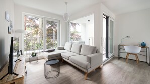 Een moderne woonkamer met een hoekbank, een ronde salontafel en een televisie op een houten standaard. Er is een balkon met een balustrade en uitzicht op bomen.