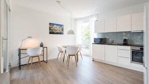 Een moderne keuken met witte kasten, een eethoek met een ronde tafel en stoelen, en een klein bureau met een lamp.