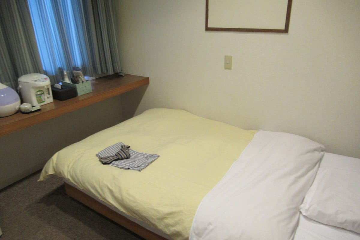 A hotel room with a bed, a window with curtains, a bedside table with a coffee maker, and a wall-mounted light switch.