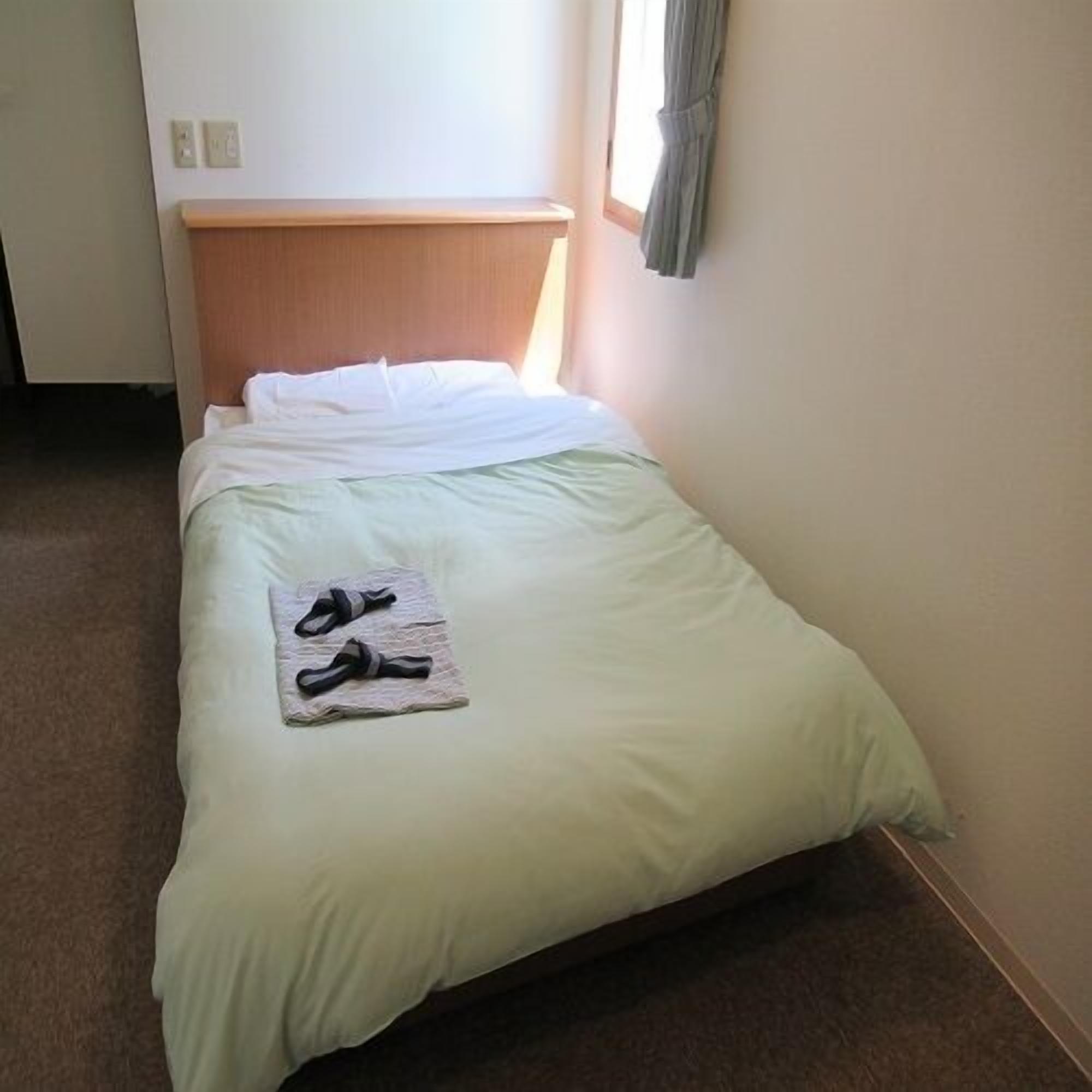 A single bed with a wooden headboard, a green bedspread, and a pair of slippers on a folded towel.