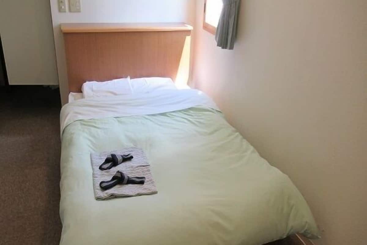 A single bed with a wooden headboard, a green bedspread, and a pair of slippers on a folded towel.