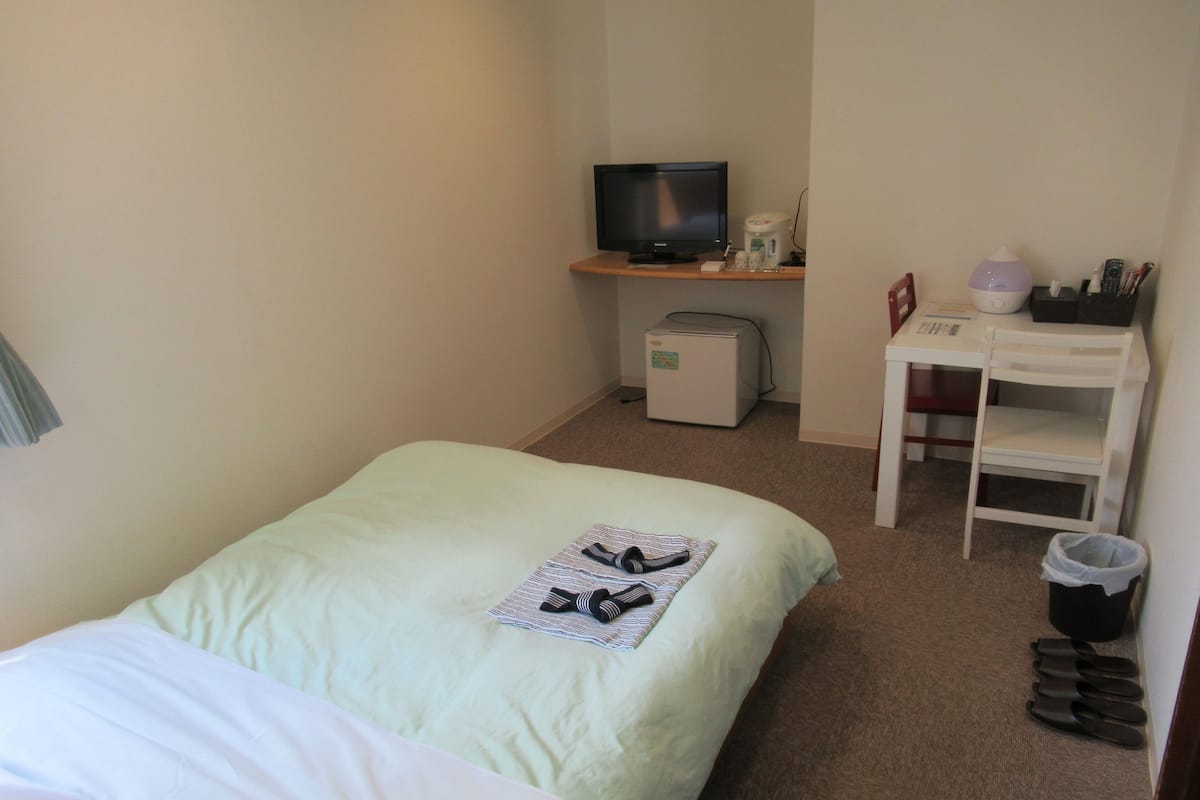 A hotel room with a bed, a desk with a computer, a television, and a trash bin.