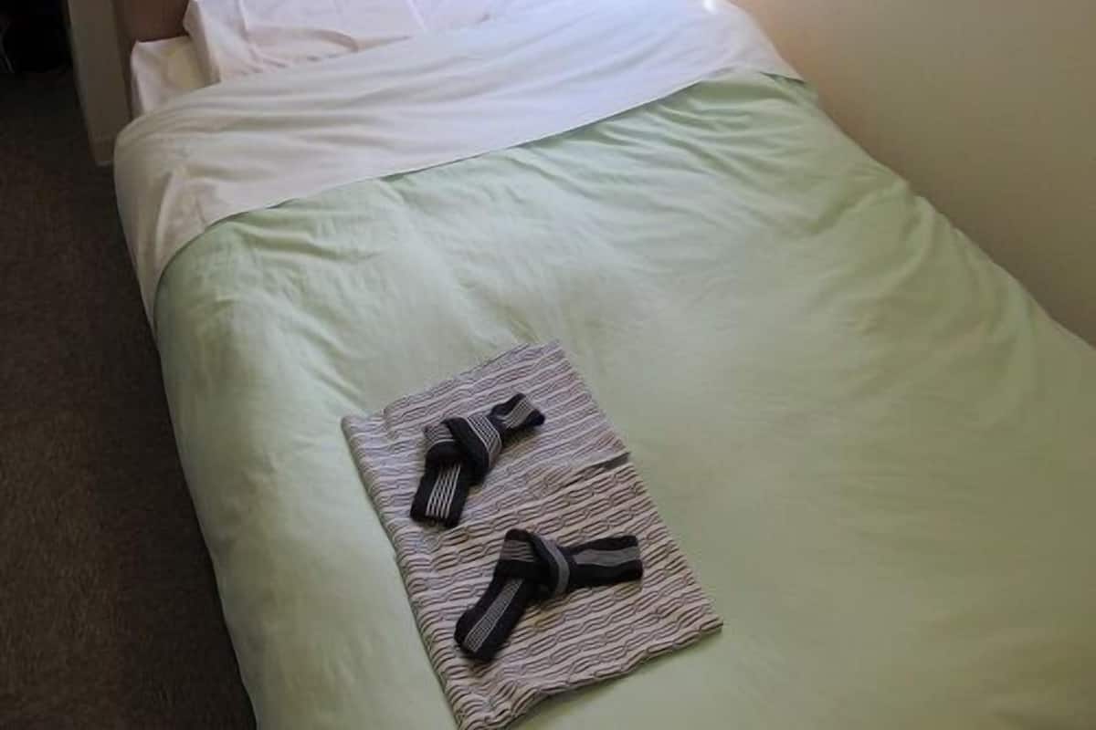 A single bed with a wooden headboard, a green bedspread, and a folded towel with black stripes on the foot of the bed.