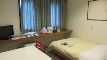 A hotel room with a single bed, a desk with a computer, a chair, a window with curtains, and a small table with a lamp and a cup.