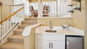 A kitchen with a sink, cabinets, and a refrigerator. There is a staircase with a handrail leading to a windowed area with chairs.
