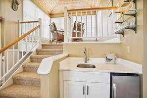 A kitchen with a sink, cabinets, and a refrigerator. There is a staircase with a handrail leading to a windowed area with chairs. - The Taverner at Ships Watch in Duck - Great Semi-Ocean Front Property (Duck)