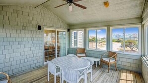 A room with a wooden floor, a ceiling fan, a table with chairs, and large windows offering a view of the outside.