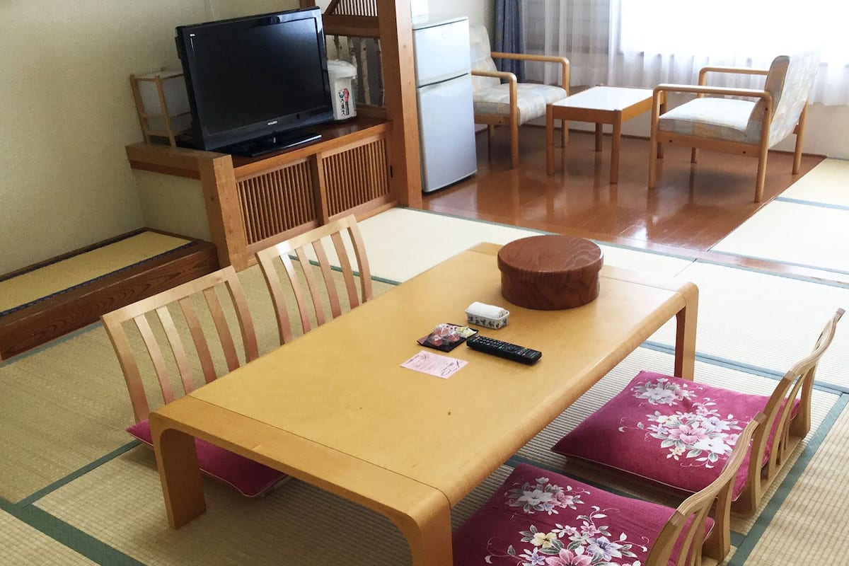 A traditional Japanese-style room with tatami flooring, a wooden table, chairs, a television, and a refrigerator.