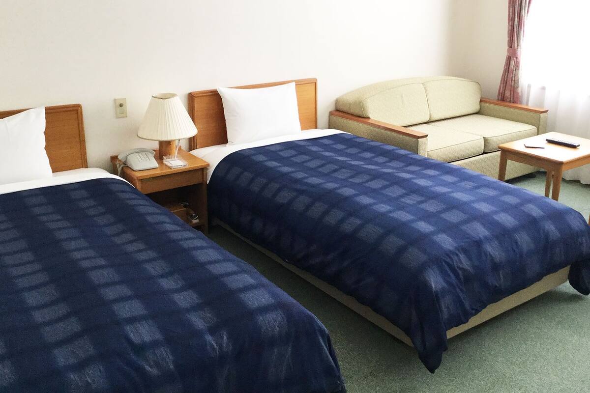 A hotel room with two beds, a chair, a small table, a lamp, and a telephone.