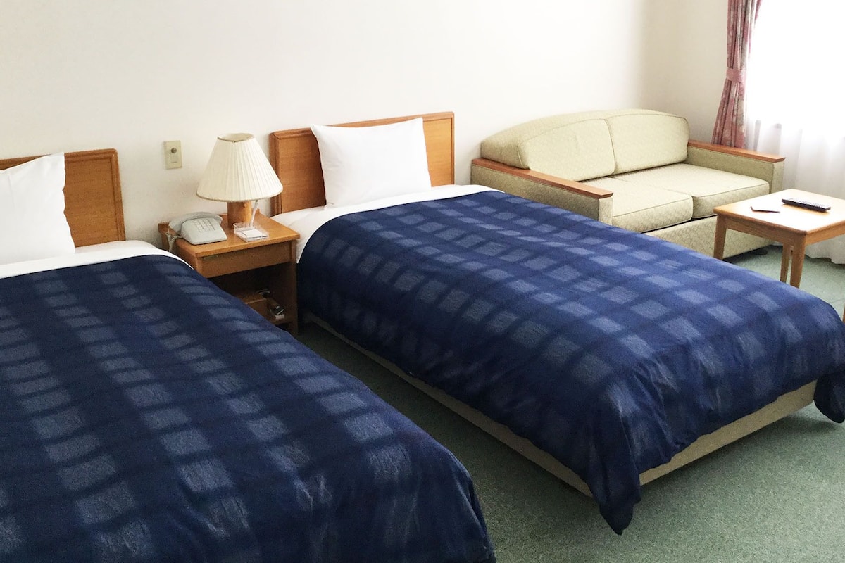 Una habitación de hotel con dos camas, una silla, una mesita pequeña, una lámpara y un teléfono.