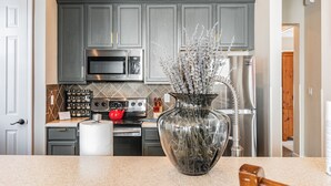 A kitchen with grey cabinets, stainless steel appliances, a large vase with dried branches, and a wooden hammer on the counter. - 2022 Award @ Hosthub, Home Theater/Game Room/Harry Potter Rm Close to Disney (Reunion)