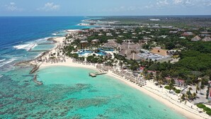 Vista aérea de un resort costero con playa, aguas turquesas cristalinas y varios edificios, incluyendo villas del resort y una zona de piscina.