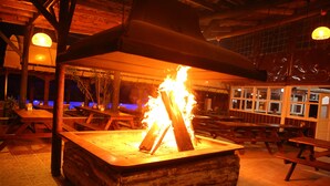 Ein hölzerner Feuerkorb mit brennenden Holzscheiten in ländlicher Umgebung, mit Holzbanken und Tischen.