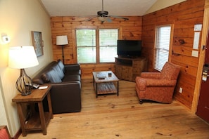 L’intérieur d’une cabane en rondins, avec un canapé, un fauteuil, une table basse, un meuble pour la télévision et un ventilateur de plafond.