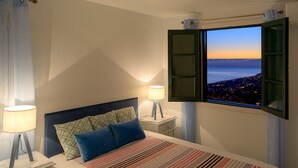 A bedroom with a bed, two bedside lamps, a window with a view of a city at dusk, and a nightstand with a lamp. - Recanto das Florenças (1) – Great Views & Sunsets (Arco Da Calheta)