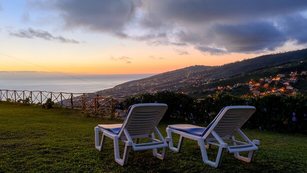 Two white lounge chairs on a grassy lawn overlooking a coastal town at sunset. - Recanto das Florenças (1) – Great Views & Sunsets (Arco Da Calheta)