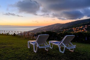 Zwei weiße Lounge-Stühle auf einer grünen Wiese mit Blick auf eine Küstenstadt bei Sonnenuntergang.