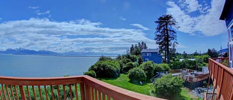 A wooden deck with a view of a lake, mountains, and a blue house with a deck.